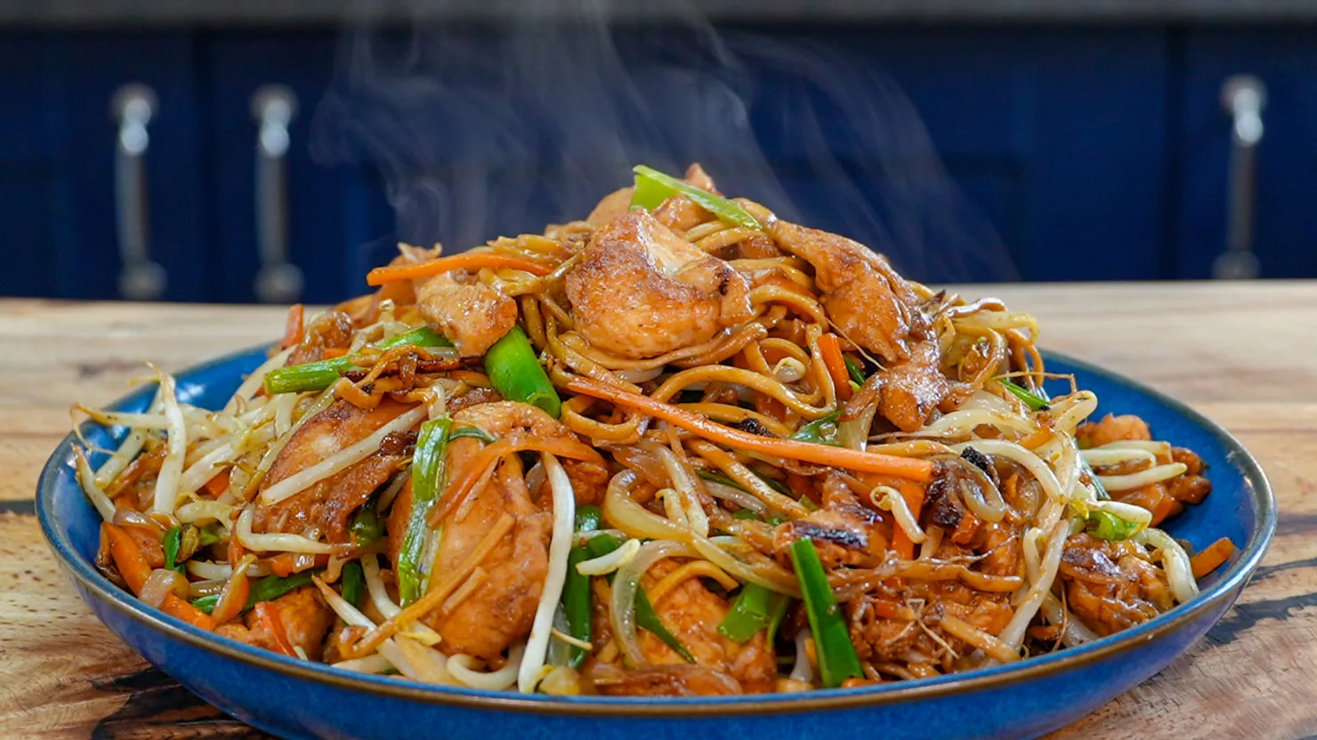 Steaming plate of stir-fried noodles with chicken, carrots, and bean sprouts on a wooden table, vibrant and appetizing.