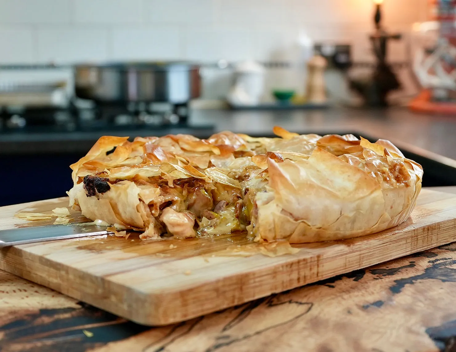 Golden-brown savory pie with crispy crust on a wooden board, a slice pulled out. Background: kitchen with pots and a soft warm light.