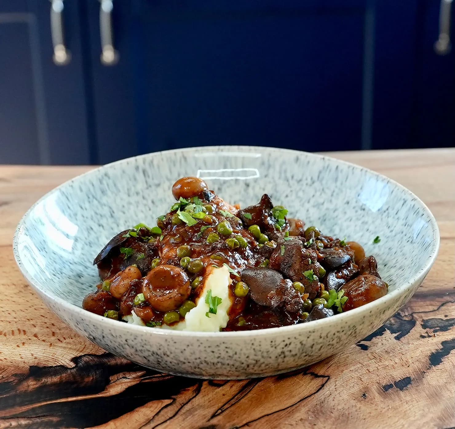 Hearty chicken liver stew with peas and mushrooms in a speckled bowl on a wooden table, set against a dark blue kitchen background.