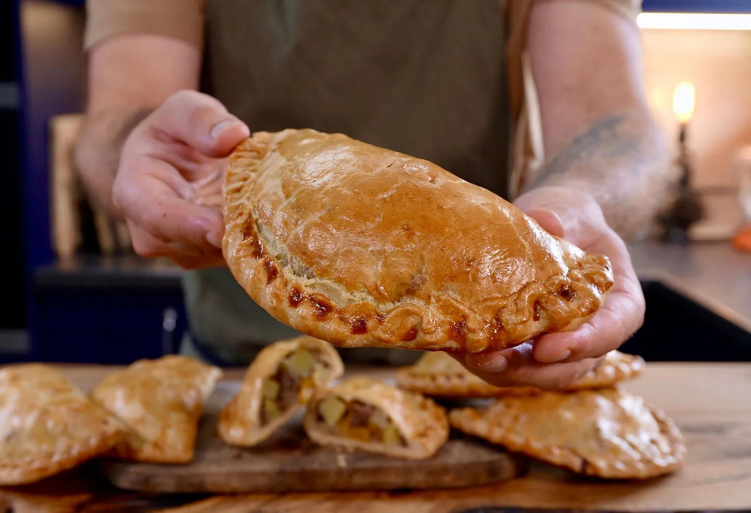 Hands holding a golden-brown Cornish pasty in a kitchen. More pasties with filling are visible on a wooden board. Warm, inviting atmosphere.