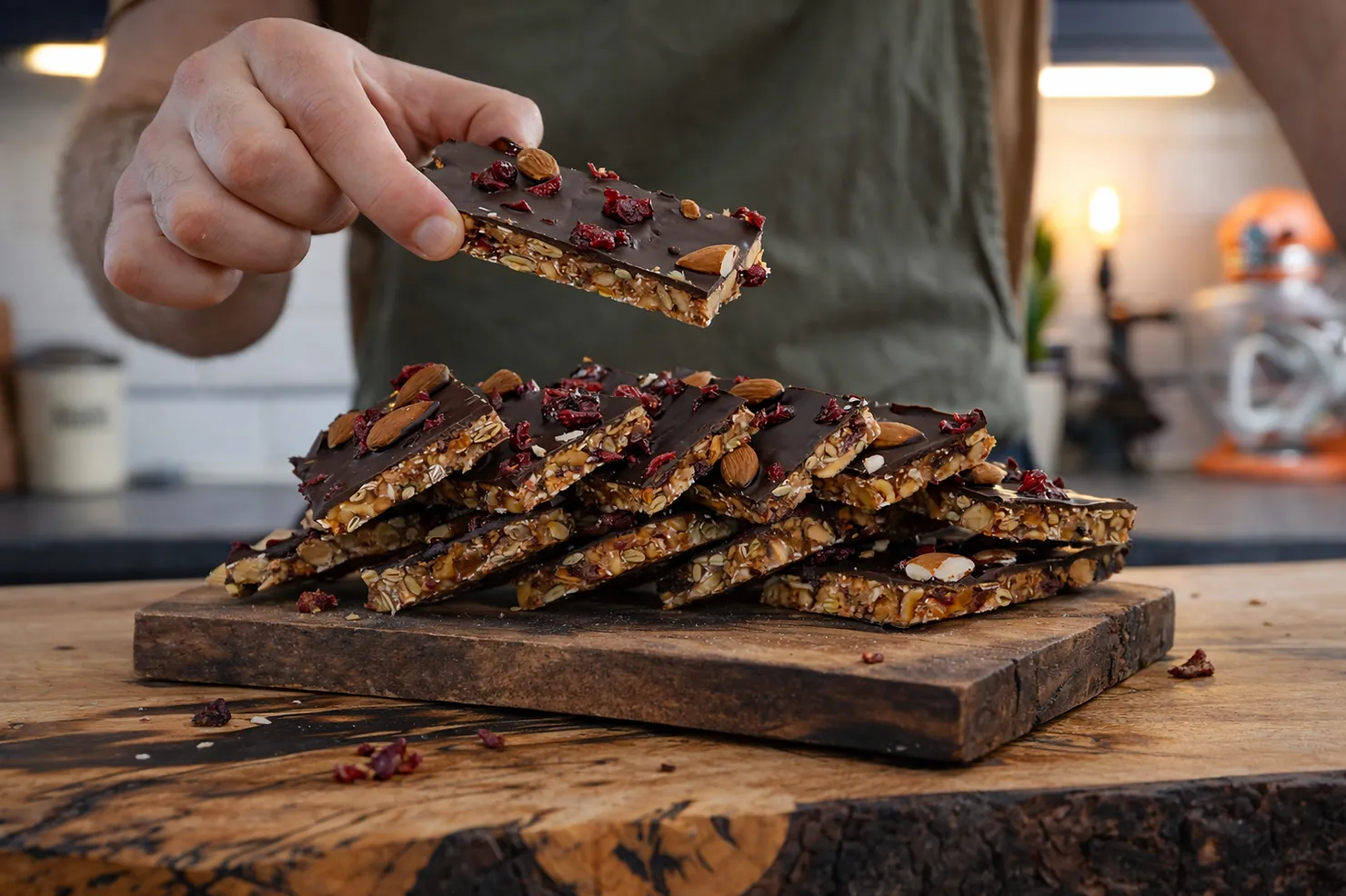 A hand holds a chocolate almond granola bar with cranberries, stacked on a wooden board in a cozy kitchen setting.