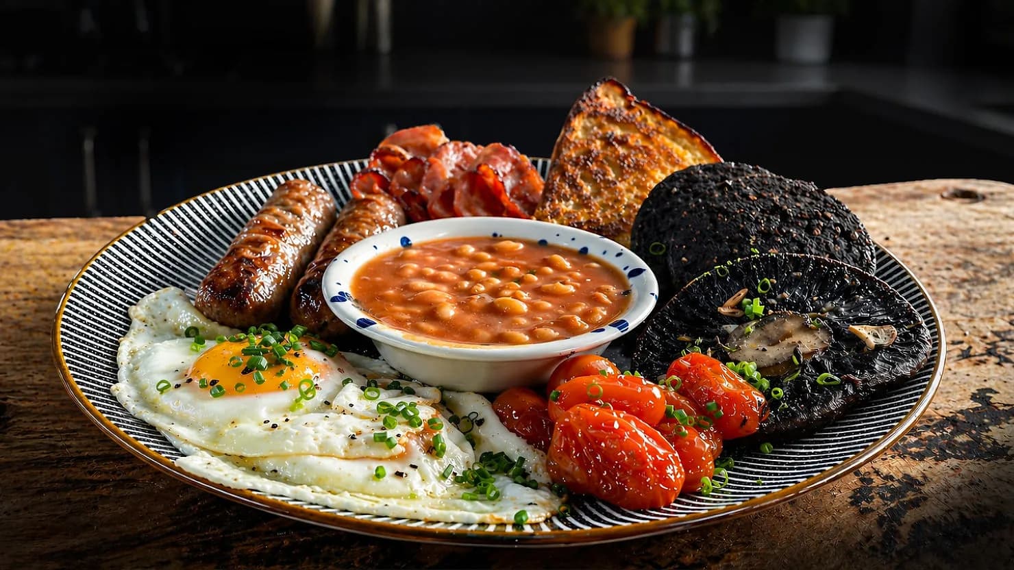 Plate of English breakfast with fried eggs, sausages, baked beans, bacon, toast, tomatoes, and black pudding on a wooden table.