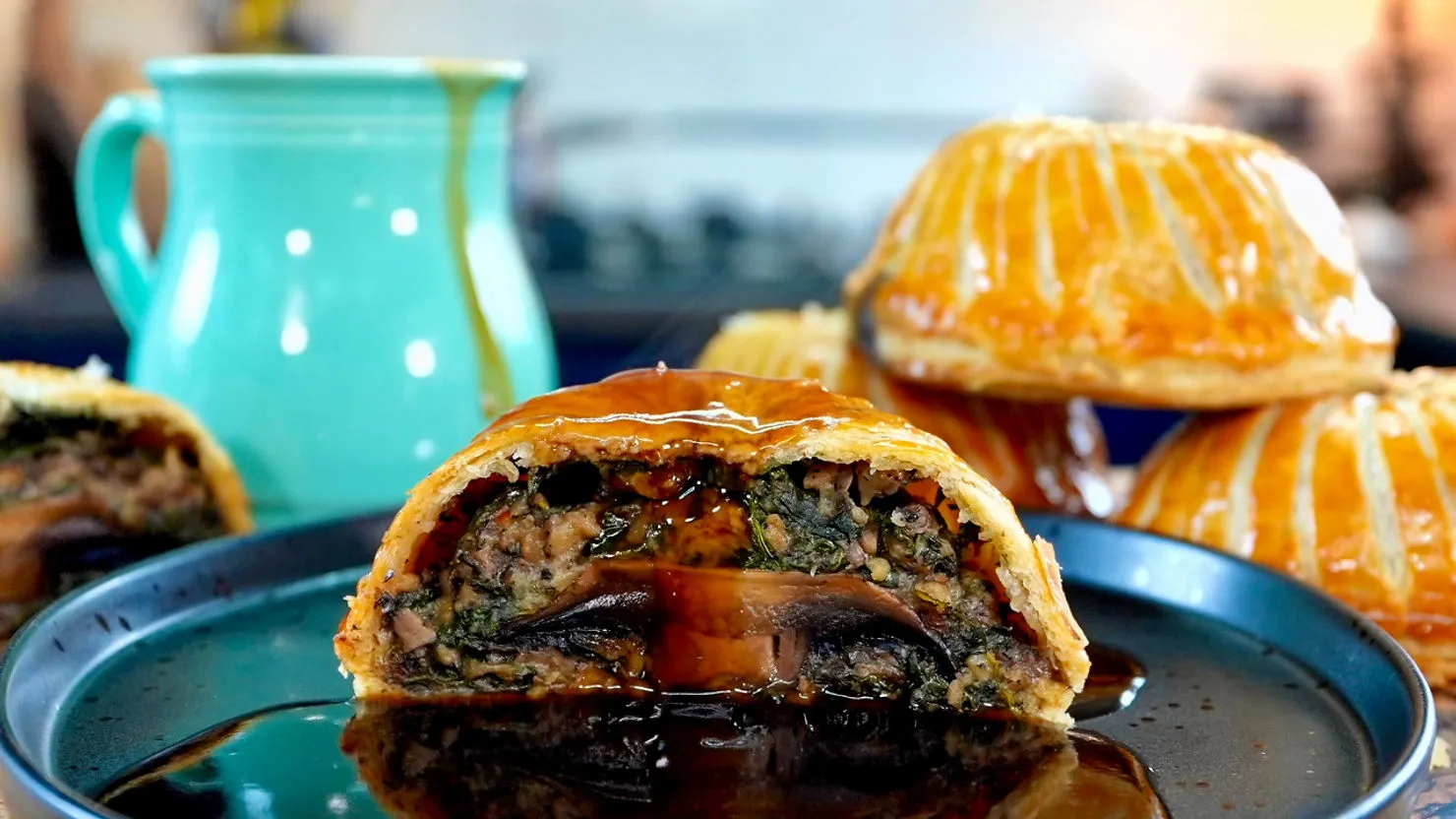 Close-up of a cross-sectioned pastry filled with mushrooms and greens, drizzled with sauce, on a black plate. Turquoise mug in background.