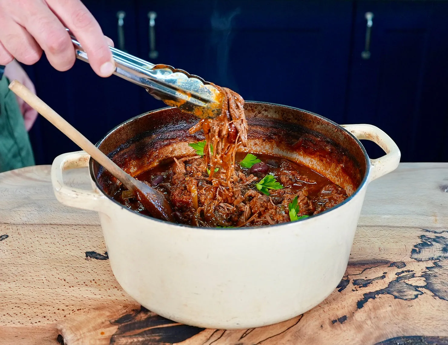 Hand using tongs to lift tender meat from a white pot containing rich, brown stew with parsley, set on a wooden table. Warm, comforting mood.