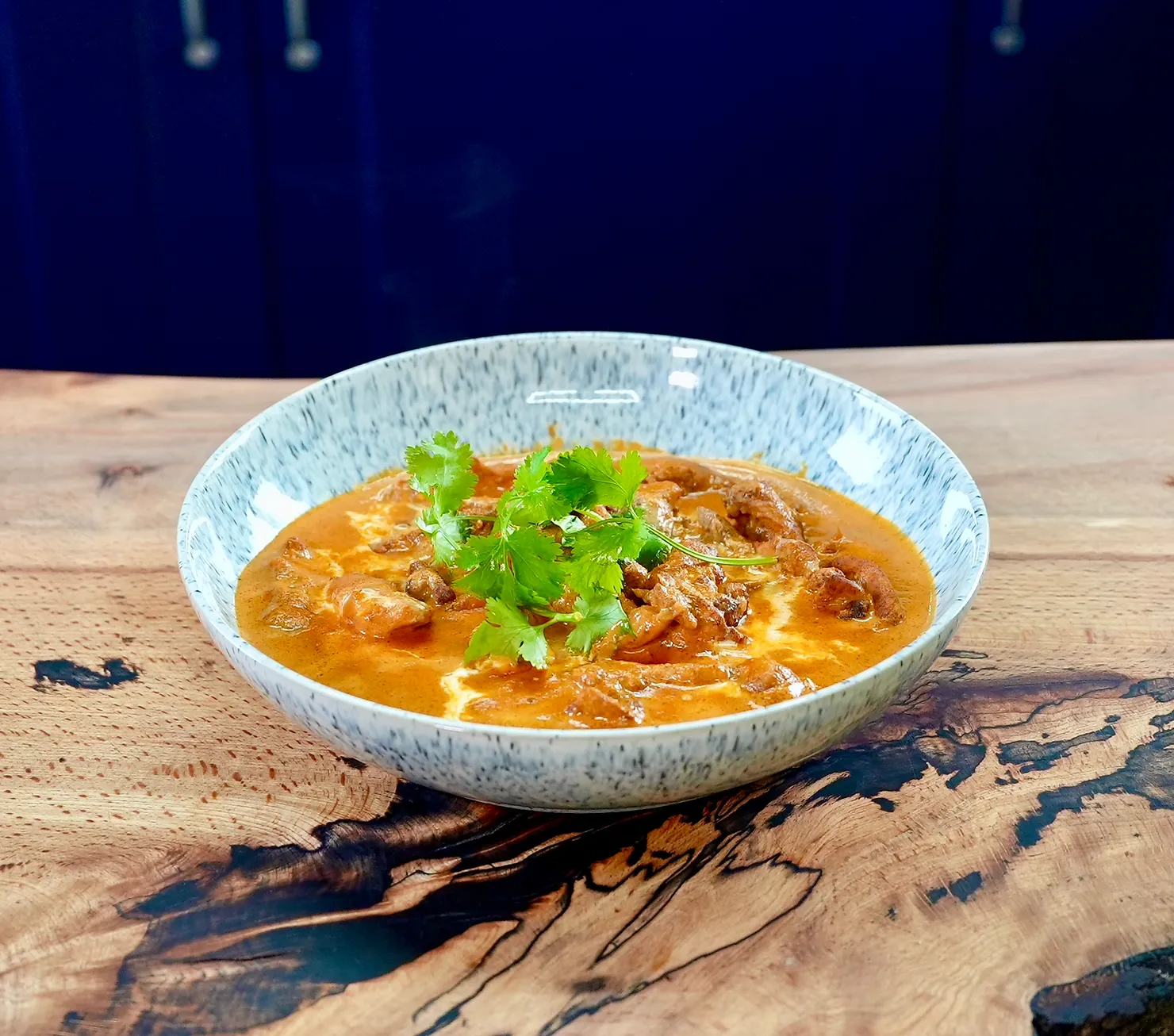 A bowl of curry topped with fresh cilantro sits on a rustic wooden table. The curry is rich orange with creamy swirls, set against a dark background.