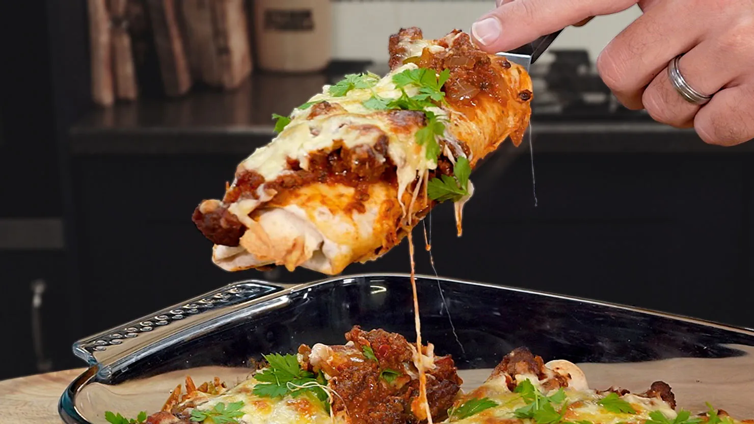 Hand lifting cheesy enchilada from a baking dish, topped with sauce and cilantro. Kitchen setting with blurred background.