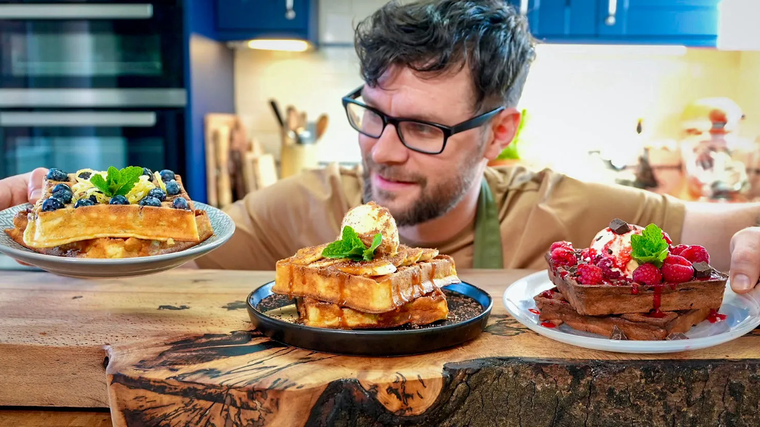 Man smiling at three plates of waffles topped with blueberries, bananas, and raspberries. Bright kitchen setting, warm mood.