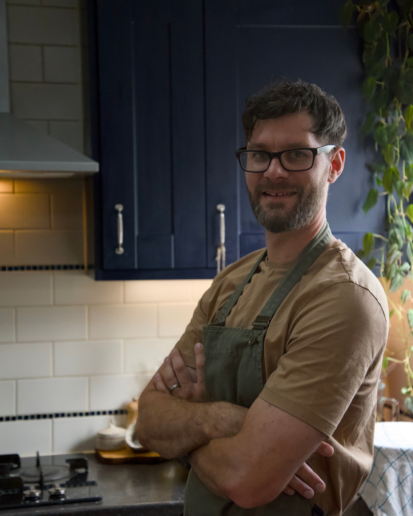 Tom standing side-on at the hob in a navy kitchen, arms folded