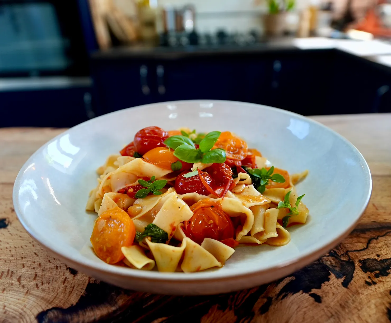 A plate of pasta topped with cherry tomatoes and basil sits on a wooden table in a cozy kitchen, creating a fresh and inviting vibe.
