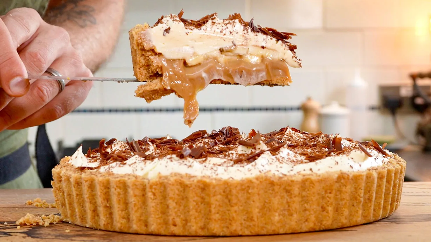 Close-up of a banoffee pie being sliced. Cream, banana, and caramel layers visible. Topped with chocolate shavings. Kitchen background.