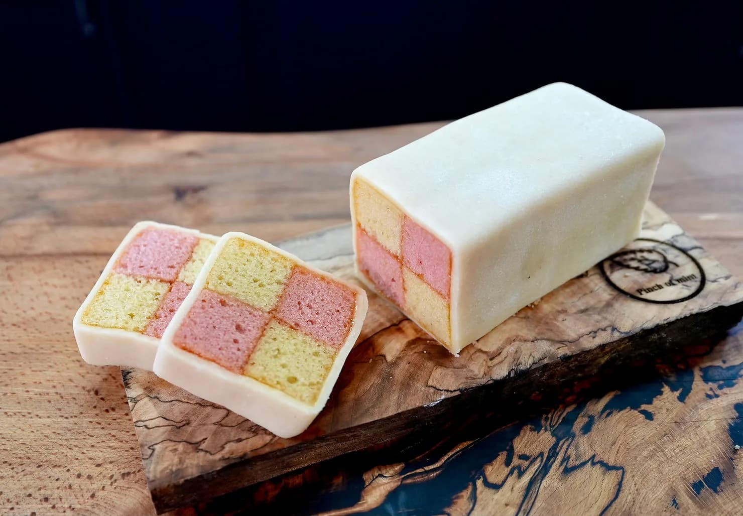 Battenberg cake on a wooden board, showing pink and yellow checkered pattern with white marzipan. Rustic kitchen setting.