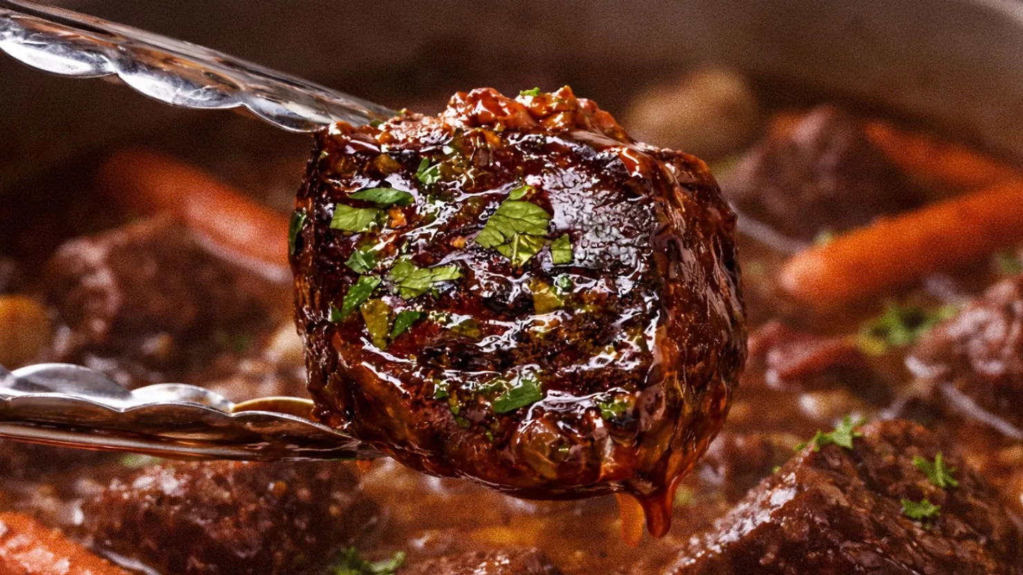 A piece of glazed, herb-garnished beef is held by tongs over a stew with carrots and meat chunks. Warm, hearty kitchen setting. Beef Bourguignon Recipe
