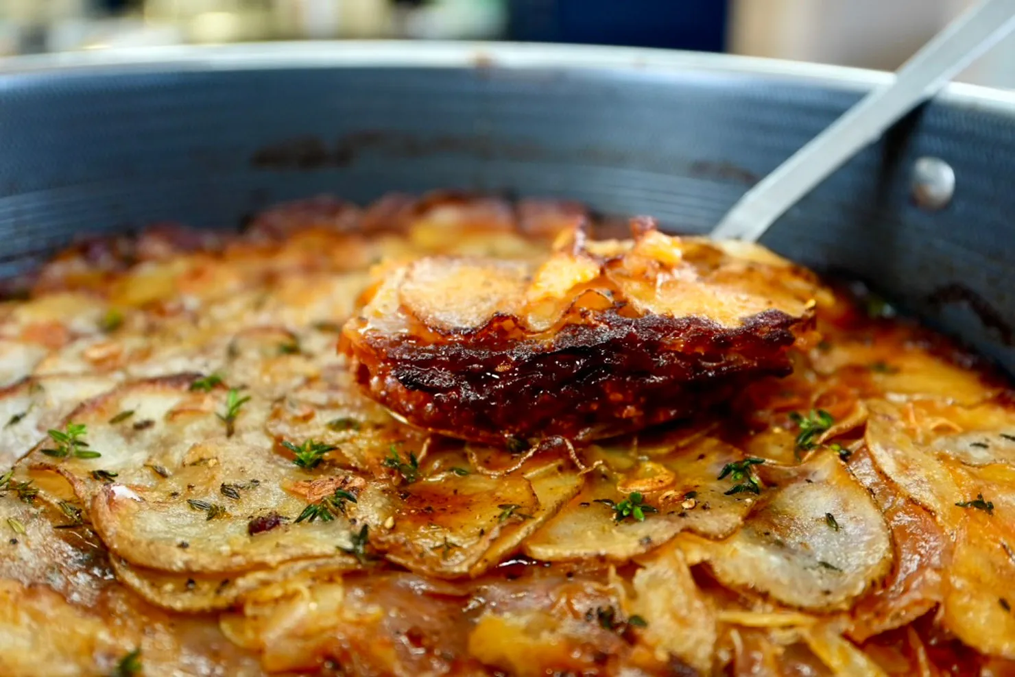 Close-up of golden-brown potatoes in a pan, with a serving spoon lifting a crispy slice. Garnished with herbs, warm and savory mood.