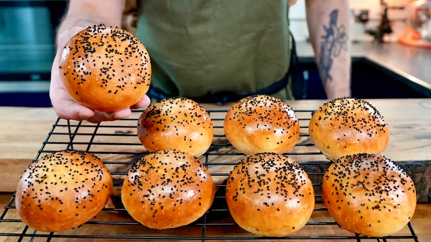 Person holding a burger bun, showcasing seven similar buns on a cooling rack in a kitchen. Bun tops are golden brown with black seeds.