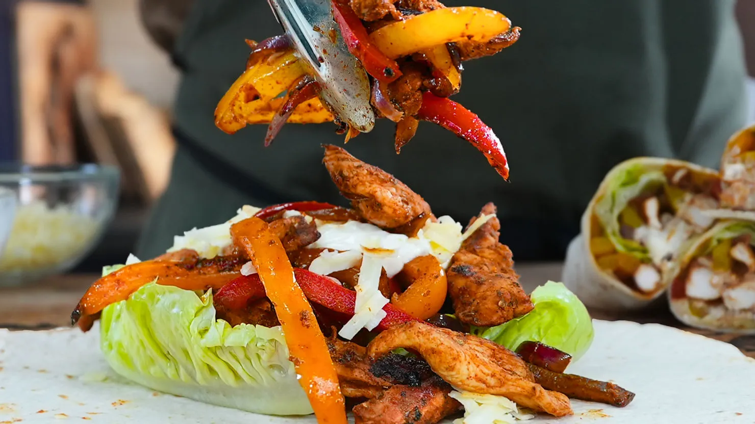 Fajita being made with grilled chicken, red and yellow peppers, lettuce, and cheese. Background shows a bowl and wrapped taco.