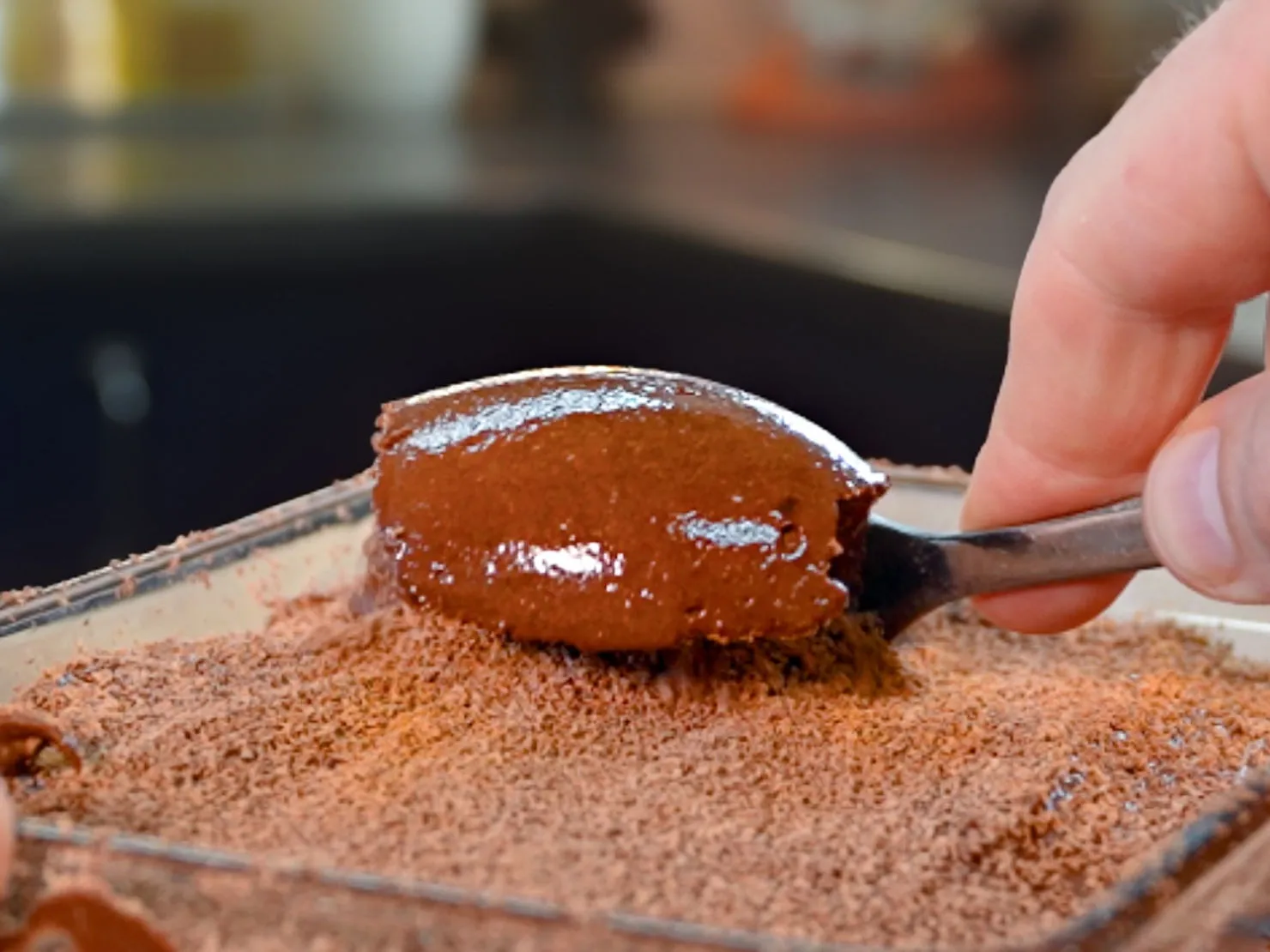 Spoon scoops shiny chocolate mousse from a glass dish, dusted with cocoa powder. Blurred kitchen background. Hands-on action.