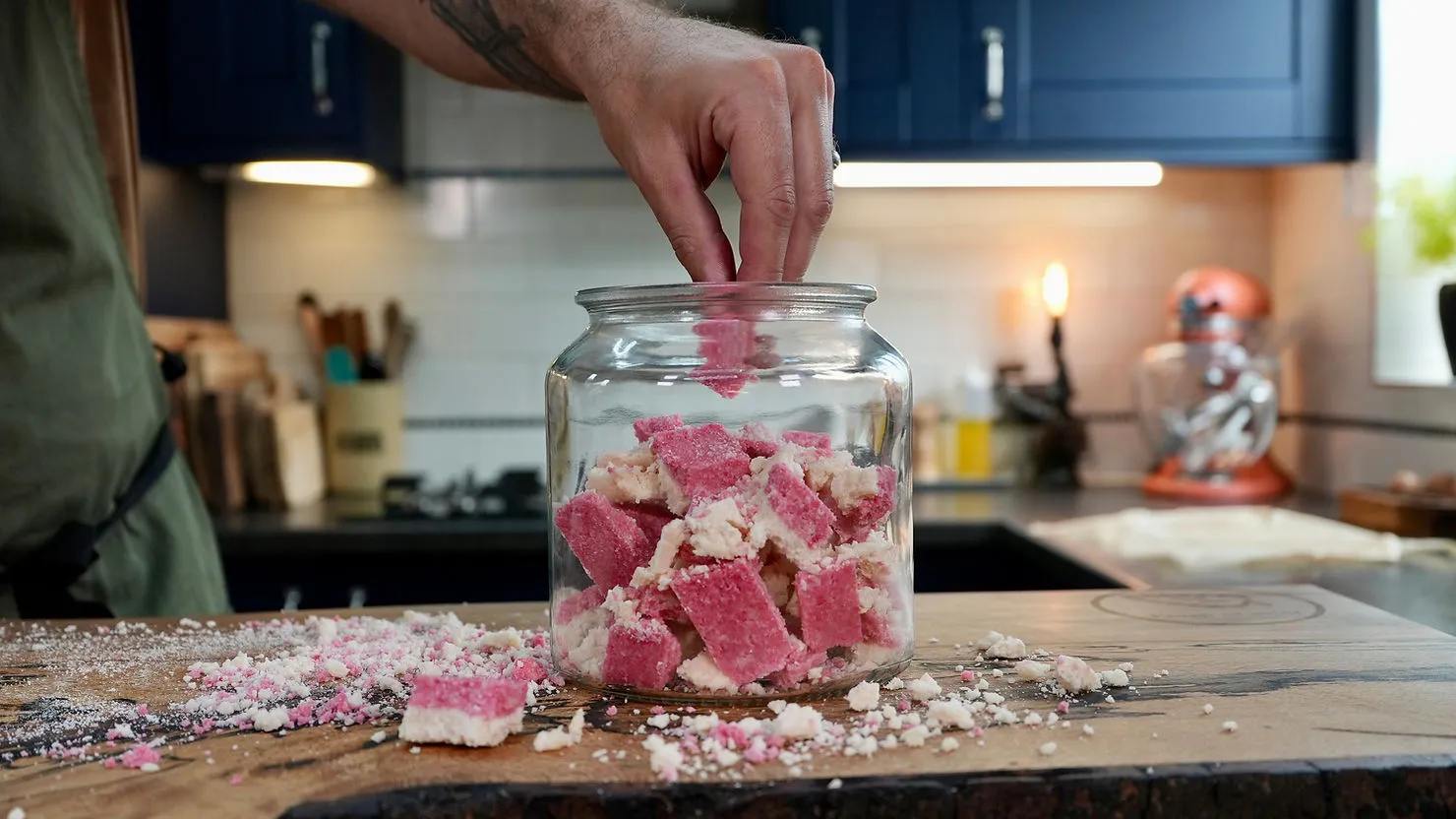 A hand places pink and white squares into a glass jar on a wooden board in a kitchen. Blue cabinets and mixer in the background.