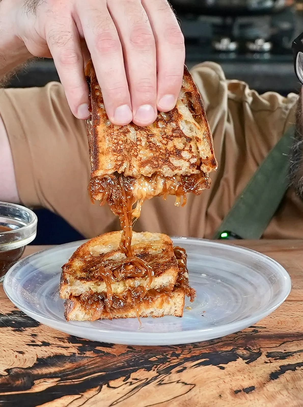 Hand pulls melted cheese from a grilled sandwich with caramelized onions on a clear plate. Wooden table, casual setting.