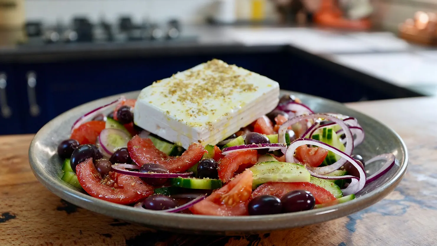 Greek salad with tomatoes, cucumbers, olives, onions, and a slab of feta, drizzled with olive oil on a wooden kitchen countertop.