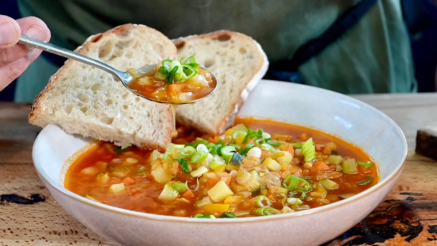 Close-up of a bowl of vegetable soup with green onions, served with slices of rustic bread. A hand holds a spoonful above the bowl.