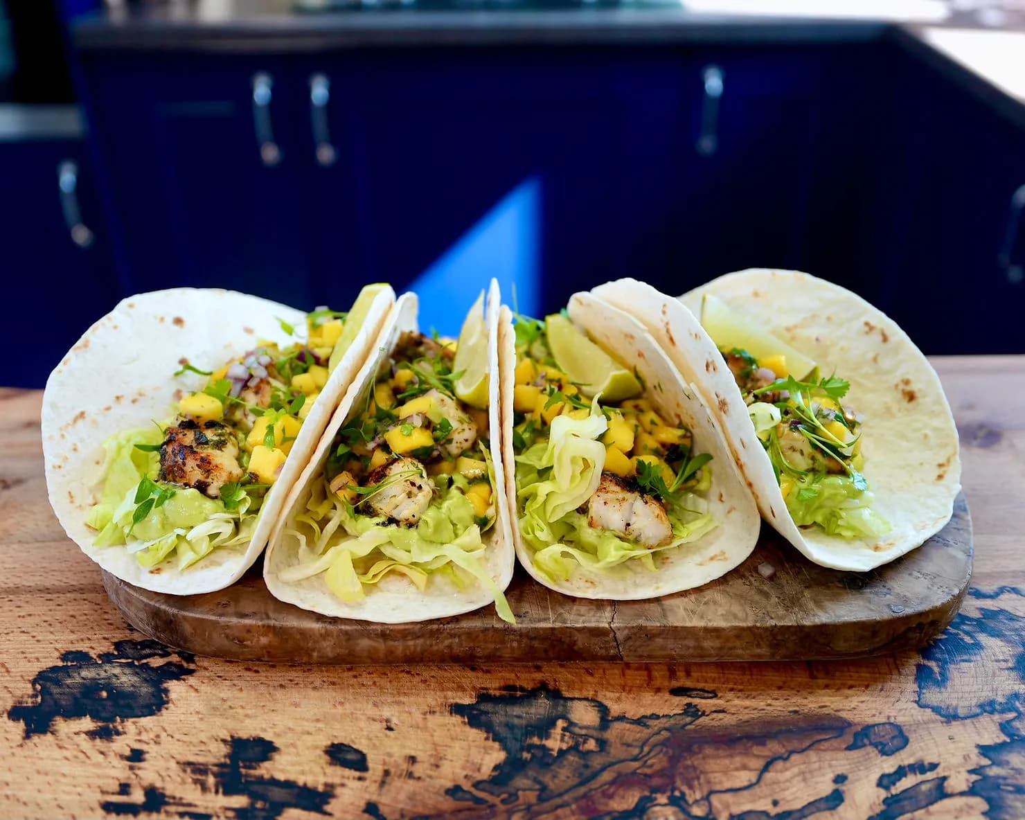 Four Monkfish tacos with lettuce, mango salsa, avocado cream and lime on a wooden board. Blue kitchen cabinets in the background.