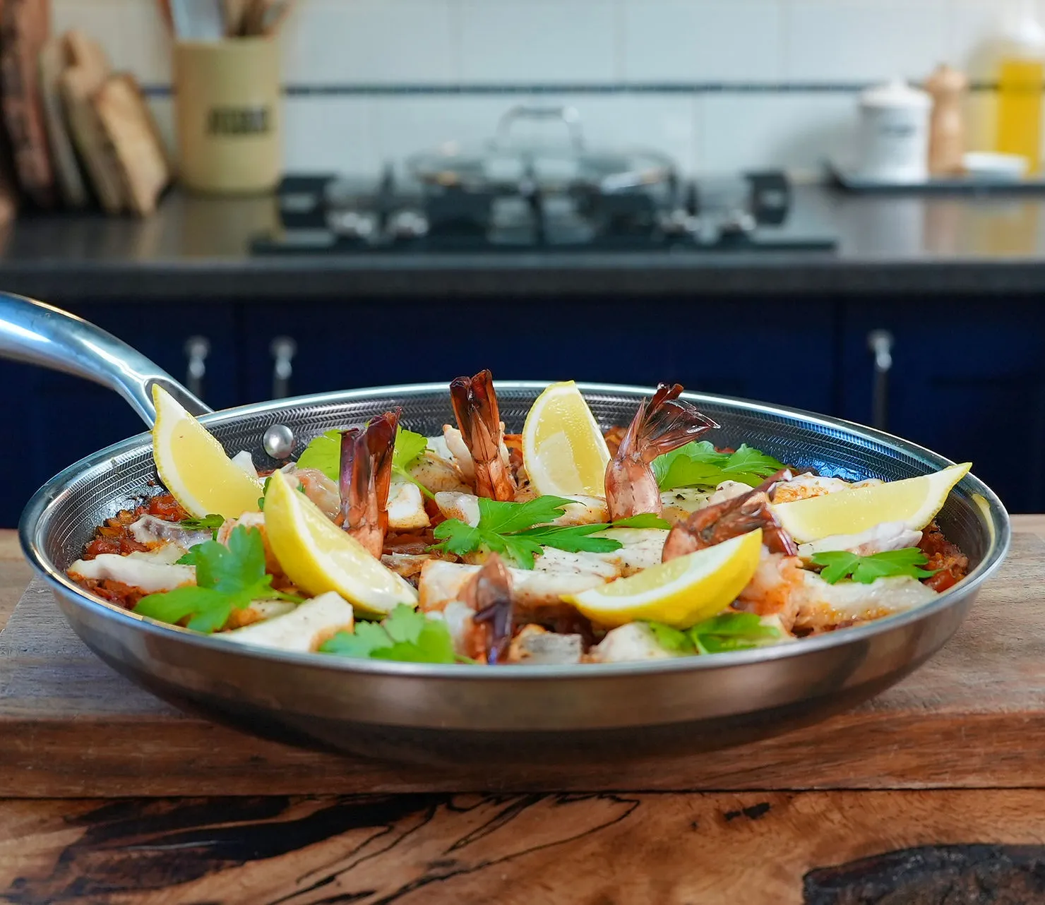 A paella pan with shrimp, lemon wedges, and parsley on a wooden surface in a kitchen setting with navy cabinets in the background.