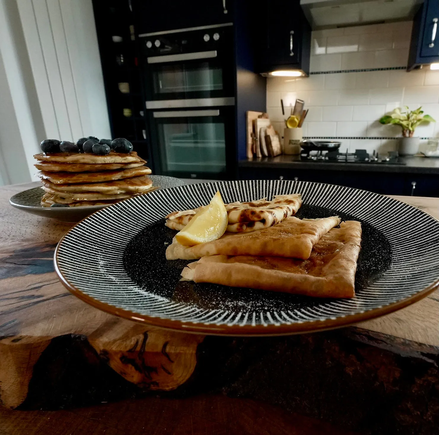 Plates of pancakes with blueberries and folded crepes with lemon in a cozy kitchen, featuring dark cabinets and wooden accents.