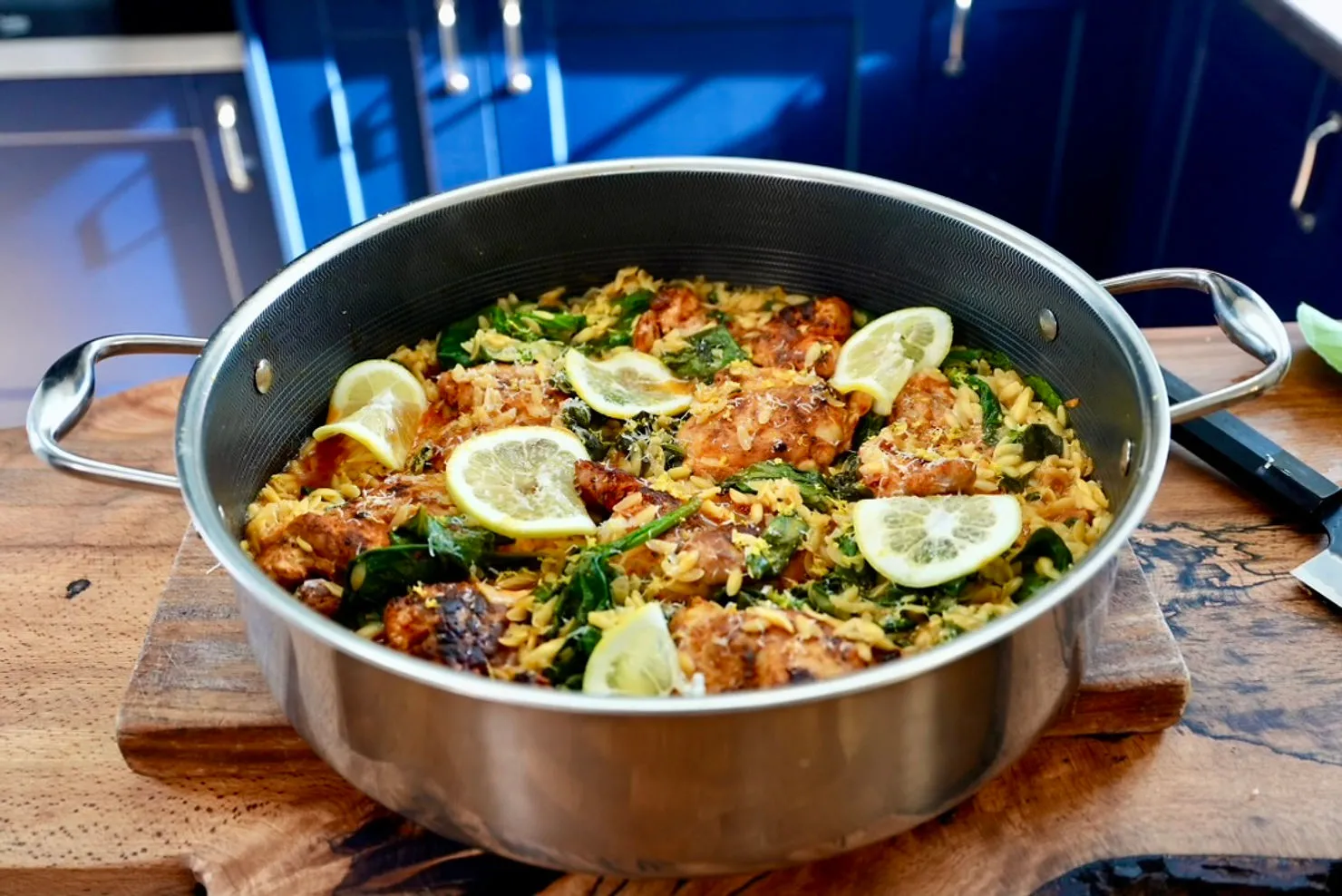 A pot of chicken, orzo, and spinach garnished with lemon slices, set on a wooden table with a blue kitchen background.