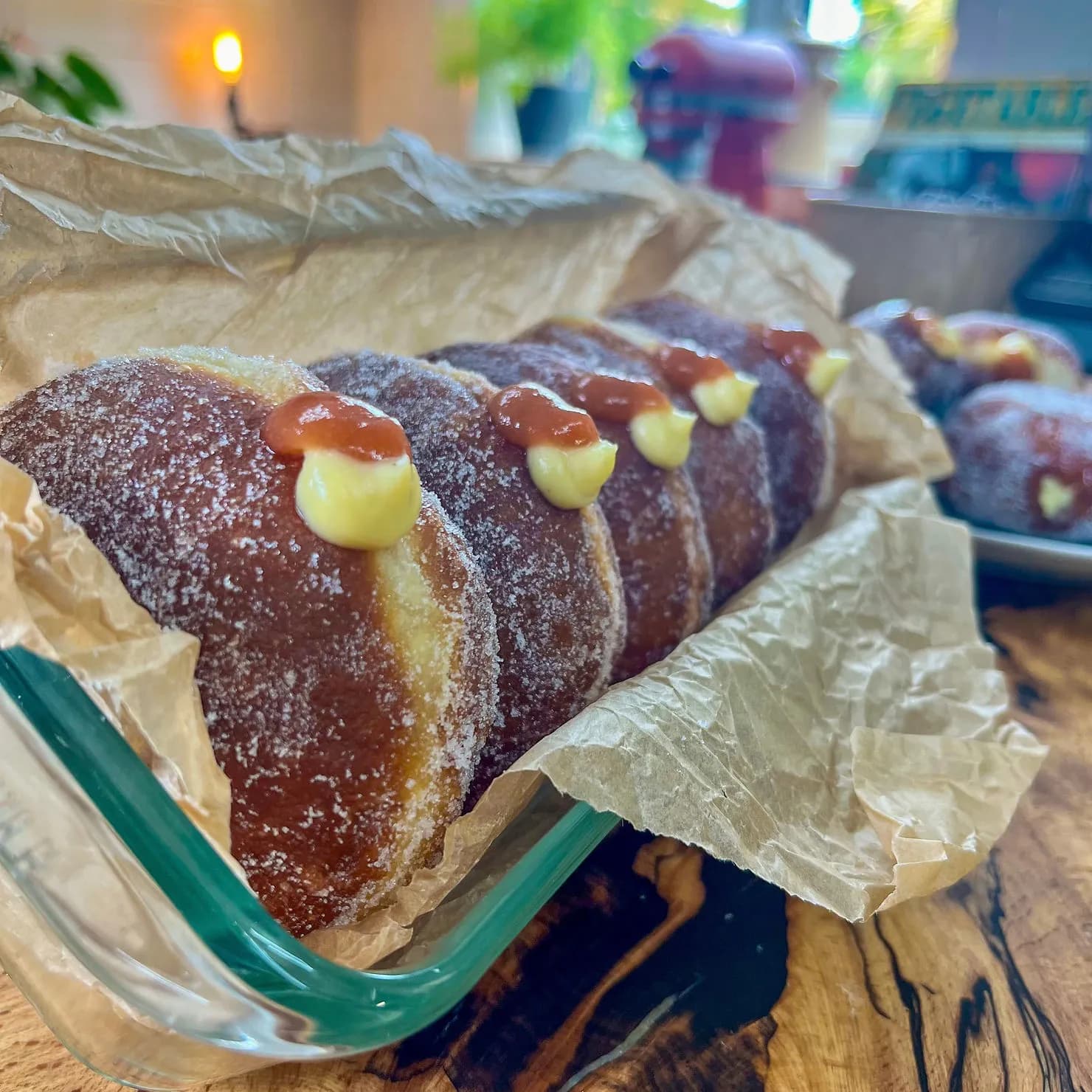Sugary donuts filled with cream and jam sit in a glass dish on a wooden table, wrapped in parchment paper. Warm kitchen background.