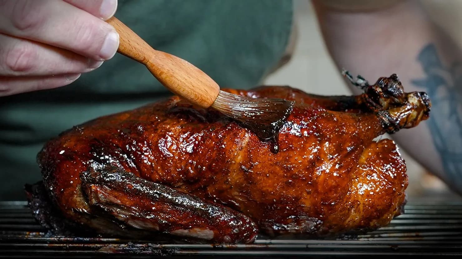 Person basting a roasted duck with sauce using a brush. The duck has a glossy brown glaze.