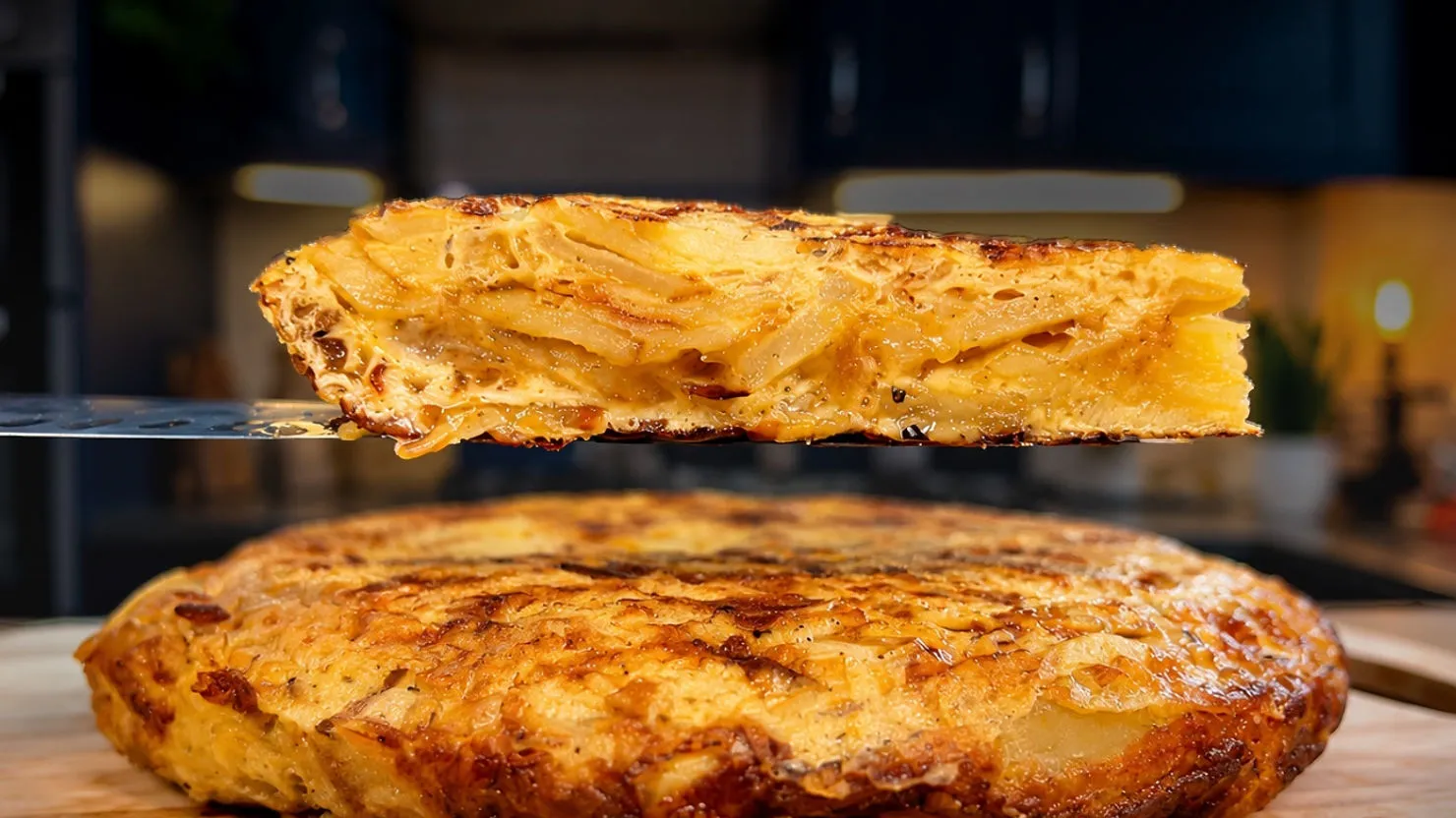 Close-up of a golden potato omelet slice on a spatula above a whole omelet on a cutting board in a blurred kitchen.