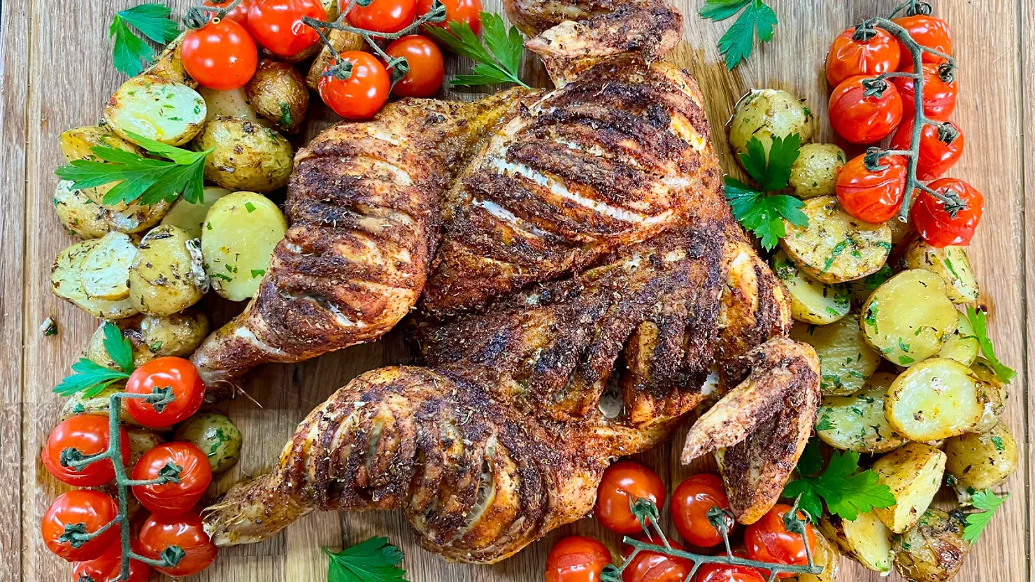 Grilled spatchcock chicken served with roasted potatoes and cherry tomatoes on a wooden board, garnished with fresh parsley.
