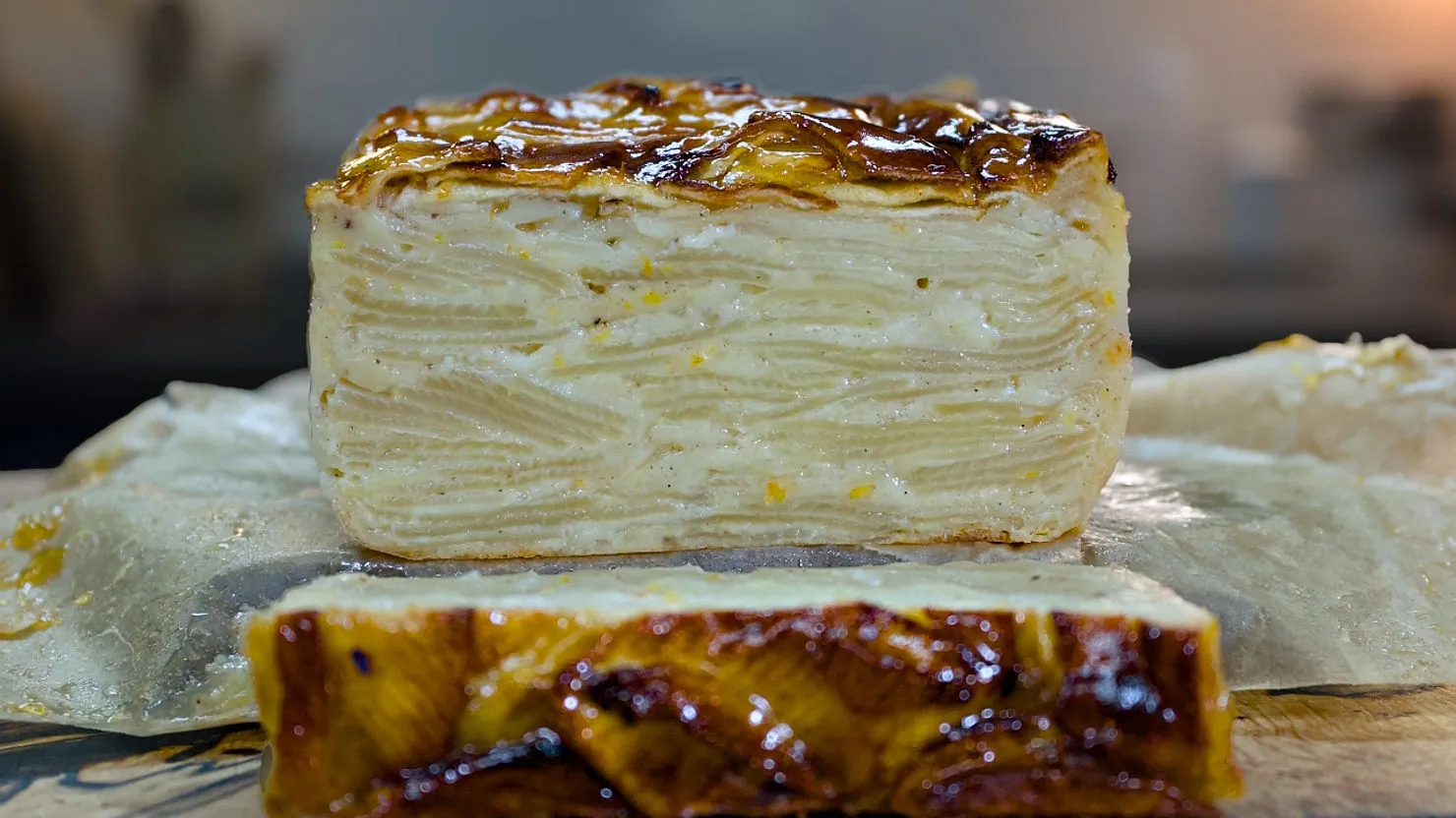 Layered apple cake on parchment paper, with a caramelized top and visible apple layers. Warm kitchen setting, soft lighting.