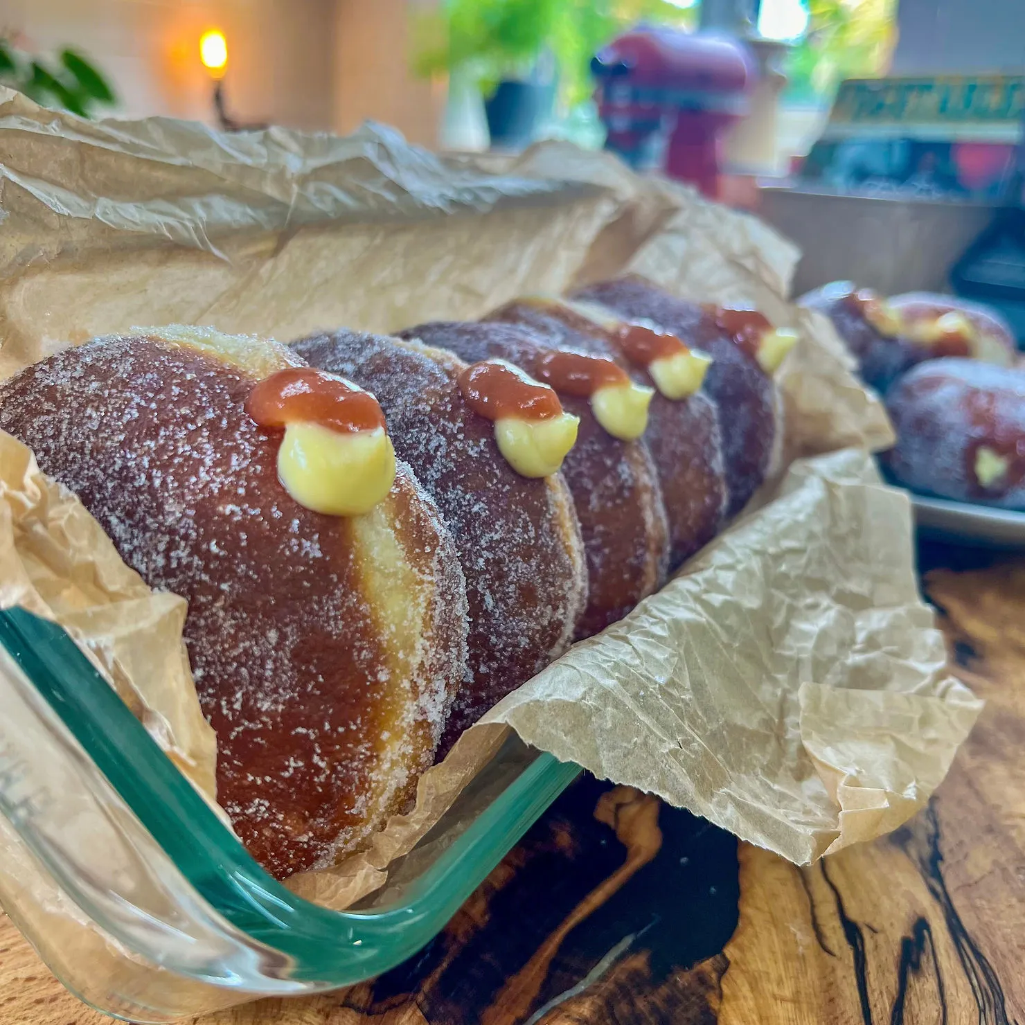 Sugary donuts filled with cream and jam sit in a glass dish on a wooden table, wrapped in parchment paper. Warm kitchen background.