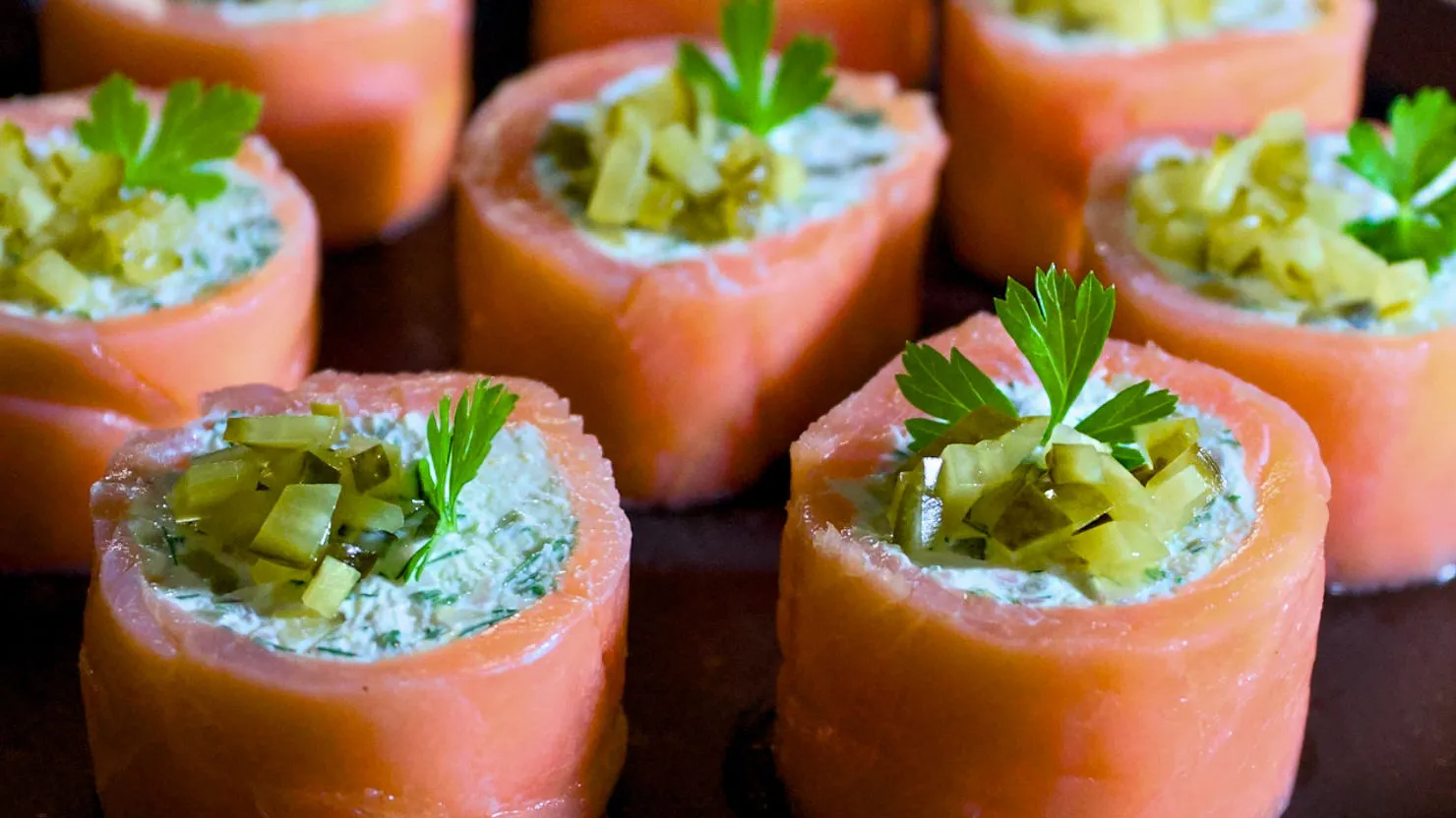 Salmon rolls filled with creamy dill, topped with pickles and parsley, arranged neatly on a dark plate. Vibrant and appetizing.