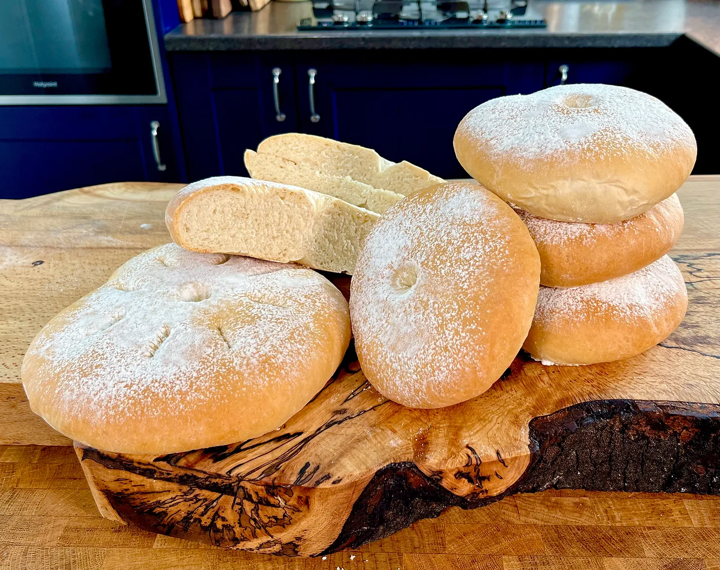 Bread rolls and sliced loaves stacked on a wooden cutting board in a dark blue kitchen.