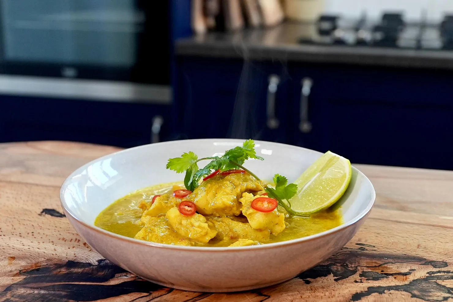 Bowl of steaming yellow curry with fish, garnished with cilantro, red chili slices, and a lime wedge. Set on a wooden countertop.